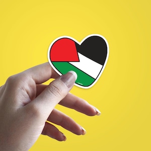 Könnte beinhalten: Ein herzförmiger Aufkleber mit dem Design der palästinensischen Flagge. Die Flagge hat die Farben Rot, Weiß, Schwarz und Grün. Der Aufkleber wird vor einem gelben Hintergrund gehalten.