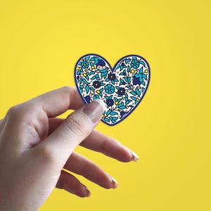 May include: A heart-shaped sticker with a floral design in shades of blue, white, and yellow, outlined in dark blue. The sticker is held against a bright yellow background. The floral pattern includes stylized flowers and leaves.