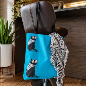 May include: A bright blue tote bag featuring a repeating pattern of black cats wearing red-rimmed sunglasses and white and black patterned scarves. A black and white fringed scarf is draped over the side of the bag, which has black fabric handles.