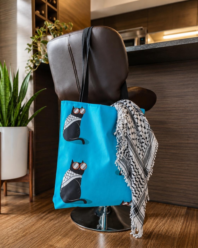 May include: A bright blue tote bag featuring a repeating pattern of black cats wearing red-rimmed sunglasses and white and black patterned scarves. A black and white fringed scarf is draped over the side of the bag, which has black fabric handles.