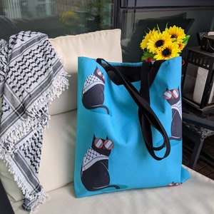 May include: A bright blue tote bag featuring a repeating pattern of black cats wearing red-rimmed sunglasses and white and black patterned scarves. The bag has two black fabric shoulder straps and is constructed from a durable, smooth textile material.