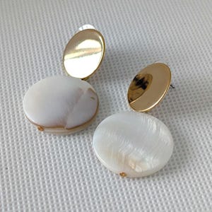 Könnte beinhalten: Ein Paar Ohrringe mit einem goldfarbenen, ovalen Stecker und einem großen, weißen, kreisförmigen Anhänger. Der Anhänger hat einen perlmuttartigen Glanz und ein subtiles, natürliches Muster. Die Ohrringe sind für durchbohrte Ohren konzipiert.