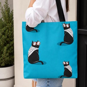 May include: A bright blue tote bag featuring a repeating pattern of black cats wearing red-rimmed sunglasses and white and black patterned scarves. The bag has two black shoulder straps and is constructed from a flat, rectangular fabric material.
