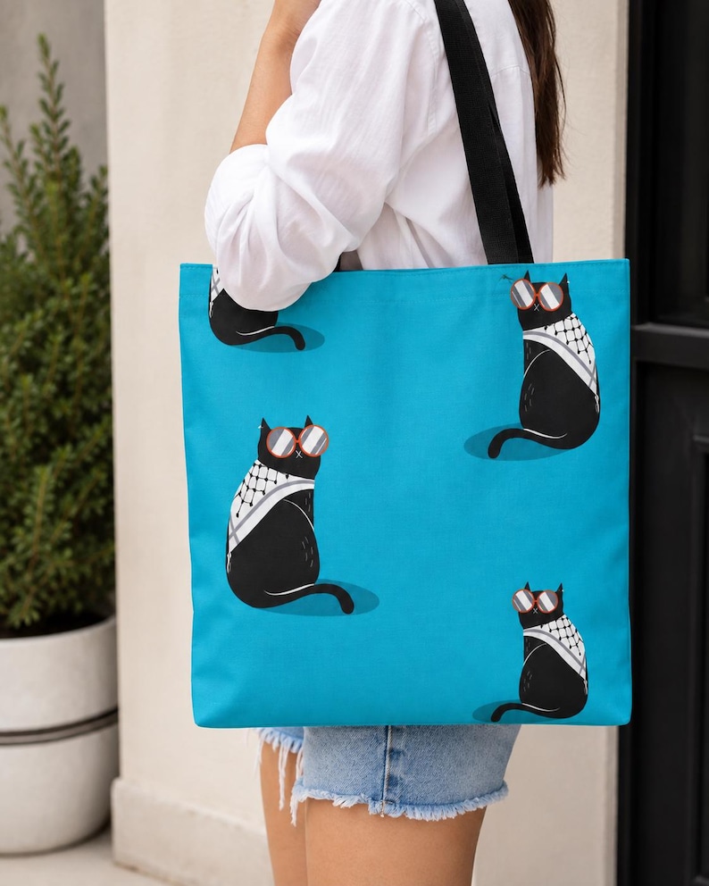 May include: A bright blue tote bag featuring a repeating pattern of black cats wearing red-rimmed sunglasses and white and black patterned scarves. The bag has two black shoulder straps and is constructed from a flat, rectangular fabric material.