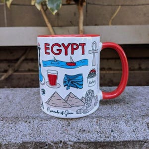 Könnte beinhalten: Weißer Keramikbecher mit rotem Innenraum und Henkel, mit dem Wort "EGYPT" und Illustrationen ägyptischer Wahrzeichen und Symbole. Der Becher zeigt Pyramiden, eine Sphinx und ein Segelboot.