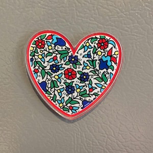 May include: Heart-shaped magnet with a red border and a floral design. The design features red, blue, and yellow flowers with green leaves on a white background. The magnet is on a gray surface.