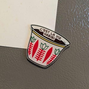 May include: A white and red coffee cup-shaped magnet with the text "C'EST LA VIE HABIBI". The cup has a yellow rim and red and green floral designs. The magnet is on a grey surface.