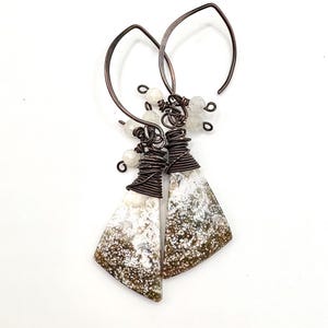 May include: Handmade earrings featuring natural stone pendants with a speckled pattern in shades of white, brown, and green. The pendants are wrapped in copper wire and suspended from copper ear wires. Small white beads add detail.