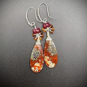 May include: A pair of handcrafted earrings featuring teardrop-shaped stones with red, orange, and gray patterns. The earrings have silver-colored ear wires and are adorned with clusters of red and amber-colored beads.