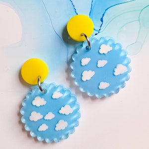 May include: A pair of blue and yellow earrings with a scalloped edge. The blue earrings feature a white cloud design. The yellow earrings are solid.
