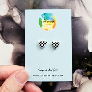 May include: Heart-shaped stud earrings with a black and white chequered pattern. The earrings are presented on a light blue card with the text "Surgical Steel Post" and the website address "www.karenhornart.co.uk".