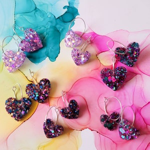 May include: Ten pairs of silver hoop earrings with heart-shaped charms. The hearts are made of glittery resin and come in a variety of colours, including pink, purple, blue, and green.