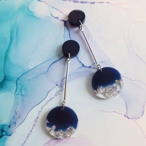 May include: A pair of dangle earrings with two round blue resin pieces with silver flakes. The top piece is smaller and the bottom piece is larger. The earrings are connected by a silver bar.