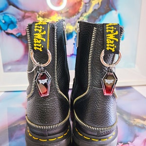 May include: A pair of black leather boots with yellow zipper pulls and silver charms. The charms are shaped like bats and have a red and black design. The boots have a black and yellow "Dr. Martens" label on the side.