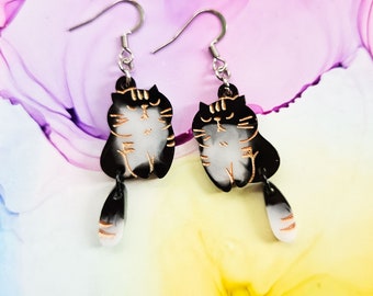 May include: A pair of dangle earrings featuring black and white cat silhouettes with gold accents. The cats have a white face and a black body with a gold outline. The earrings have a silver hook.