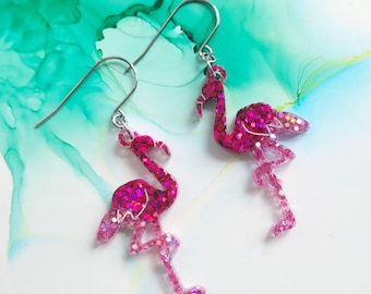 May include: A pair of dangle earrings featuring pink flamingo-shaped charms filled with vibrant magenta glitter. The flamingos are suspended from silver-toned metal hooks, showcasing a detailed silhouette with long legs and curved necks.
