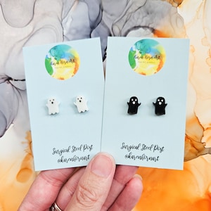 May include: Two pairs of ghost stud earrings. One pair is white and the other pair is black. The earrings are on a blue background with the text "Karen Horn Art" and "Surgical Steel Post @karen hornart".