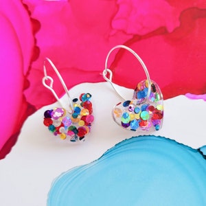 May include: A pair of silver hoop earrings with heart-shaped charms filled with colourful glitter. The glitter is in various sizes and colours, including red, blue, green, yellow, and gold.