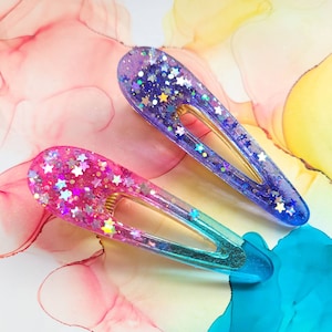 May include: Two iridescent hair clips with a gradient of pink, blue, and purple. The clips are decorated with glitter and small stars.