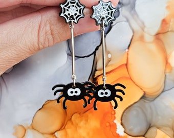 May include: A pair of black and white spider earrings with a dangling spider charm and a web design. The earrings are on a silver post.