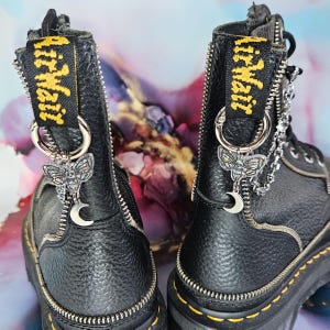 May include: Pair of black leather boots with yellow stitching and zips. Each boot features a yellow and black tag reading "Air Wair". Silver charms include a moth, a crescent moon, and a chain.