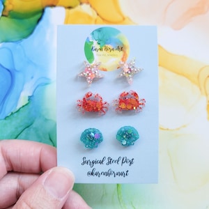 May include: A set of six stud earrings featuring a variety of sea-themed designs. The earrings are made with glitter and resin and have surgical steel posts. The designs include two starfish, two crabs, and two seashells.