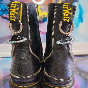 May include: Black leather boots with yellow zipper accents and a silver charm with a planet design. The charm has a glittery finish and is attached to a silver ring.