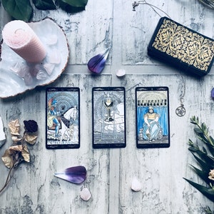 May include: A tarot card reading with three cards displayed, featuring illustrations in shades of blue, white, and gold. A deck of cards with gold filigree is visible, along with crystals, a candle, and floral accents. The cards are on a weathered, light-colored wooden surface.