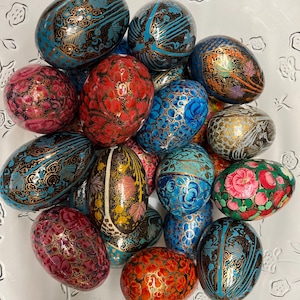 May include: A collection of oval-shaped decorative eggs featuring intricate hand-painted floral and geometric patterns in vibrant shades of red, blue, pink, and gold, arranged together in a clear glass bowl with an embossed floral design.