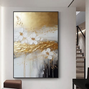 以下が含まれることがあります： 金色と白の背景に白い花が描かれた抽象画。絵画は質感があり、黒い額縁で飾られています。