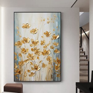 May include: A framed floral painting featuring gold-colored, three-dimensional flowers against a backdrop of cream, blue, and brown hues. The artwork has a textured appearance, with the flowers appearing to be raised from the canvas. The painting is displayed in a modern interior setting.