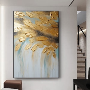 May include: Abstract painting with gold and blue paint on a white canvas. The gold paint is in the shape of leaves and the blue paint is in the shape of streaks. The painting is framed in black.