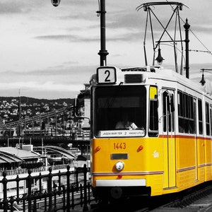 K&ouml;nnte beinhalten: Eine gelb-wei&szlig;e Stra&szlig;enbahn, Nummer 1443, f&auml;hrt auf Gleisen in einer Stadt. Die Stra&szlig;enbahn hat die Nummer 2 auf der Vorderseite. Der Hintergrund ist in Schwarzwei&szlig; gehalten, mit einer Br&uuml;cke und Geb&auml;uden.