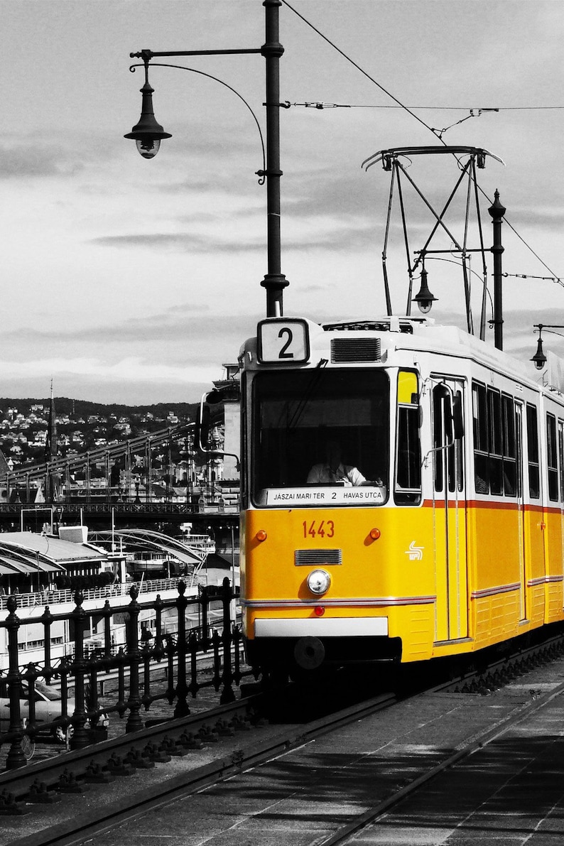 K&ouml;nnte beinhalten: Eine gelb-wei&szlig;e Stra&szlig;enbahn, Nummer 1443, f&auml;hrt auf Gleisen in einer Stadt. Die Stra&szlig;enbahn hat die Nummer 2 auf der Vorderseite. Der Hintergrund ist in Schwarzwei&szlig; gehalten, mit einer Br&uuml;cke und Geb&auml;uden.