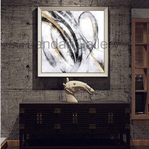Pu&ograve; includere: Opera d'arte astratta in cornice bianca, con pennellate nere e dorate su sfondo bianco. Un mobile in legno scuro con ferramenta in ottone e una scultura a testa di cavallo in pietra completano l'arredamento. L'opera &egrave; esposta su una parete grigia strutturata.