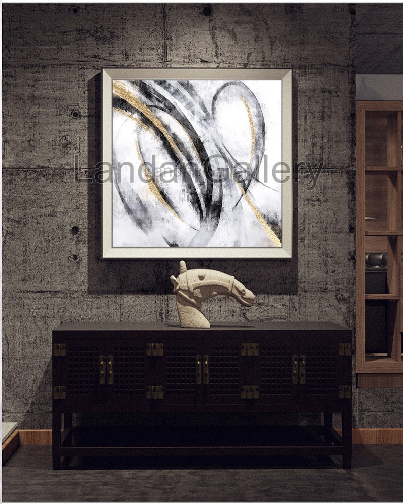 Pu&ograve; includere: Opera d'arte astratta in cornice bianca, con pennellate nere e dorate su sfondo bianco. Un mobile in legno scuro con ferramenta in ottone e una scultura a testa di cavallo in pietra completano l'arredamento. L'opera &egrave; esposta su una parete grigia strutturata.
