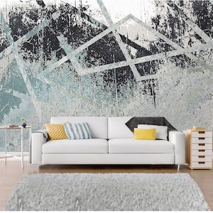 May include: A white sofa with two cushions, one yellow and one striped, sits in front of a wall with a blue and grey abstract geometric pattern. A small side table and a wooden drawer unit are also in the room.