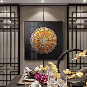 May include: A black framed canvas print featuring a gold and silver mandala design. The mandala is a circular pattern with intricate details. The print is hanging on a wall in a dining room setting with a table laid for dinner.