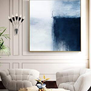 May include: Abstract painting with a white and blue color scheme. The painting is framed in gold and hangs on a white wall above two white chairs.