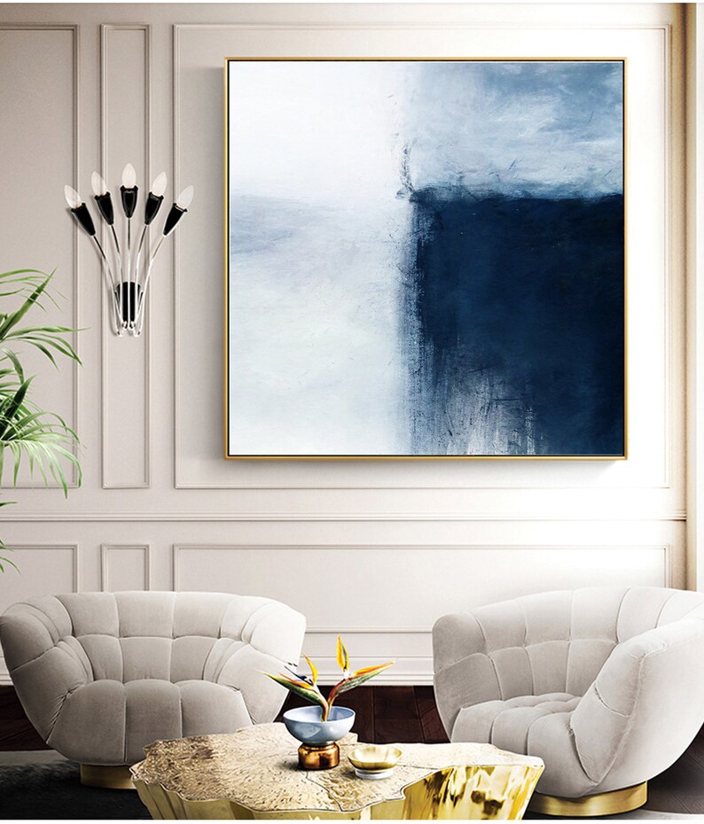 May include: Abstract painting with a white and blue color scheme. The painting is framed in gold and hangs on a white wall above two white chairs.