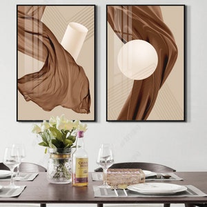 K&ouml;nnte beinhalten: Zwei abstrakte Kunstdrucke mit braunem Stoff und geometrischen Formen auf beigefarbenem Hintergrund. Die Drucke sind in Schwarz gerahmt und an einer wei&szlig;en Wand &uuml;ber einem Tisch mit einer Vase mit wei&szlig;en Blumen, einer Flasche Alkohol und Gl&auml;sern aufgeh&auml;ngt.