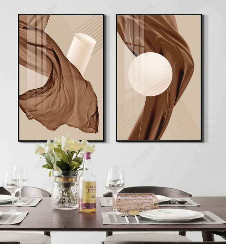 K&ouml;nnte beinhalten: Zwei abstrakte Kunstdrucke mit braunem Stoff und geometrischen Formen auf beigefarbenem Hintergrund. Die Drucke sind in Schwarz gerahmt und an einer wei&szlig;en Wand &uuml;ber einem Tisch mit einer Vase mit wei&szlig;en Blumen, einer Flasche Alkohol und Gl&auml;sern aufgeh&auml;ngt.