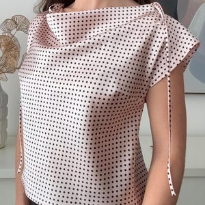 May include: A light pink top with a cowl neckline and short sleeves. The top is covered in small black polka dots. The sleeves are tied with long, thin ribbons. The top is made of a lightweight fabric.