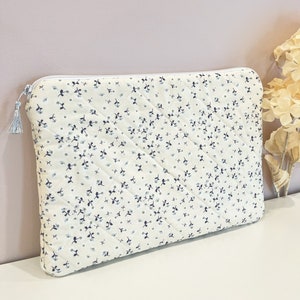 May include: A white quilted laptop sleeve with a blue floral pattern. The sleeve has a zipper closure and a tassel.