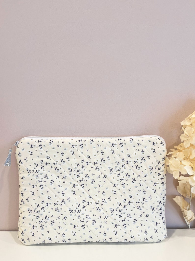 May include: A white quilted laptop sleeve with a small blue floral pattern. The sleeve has a zipper closure and a tassel pull.