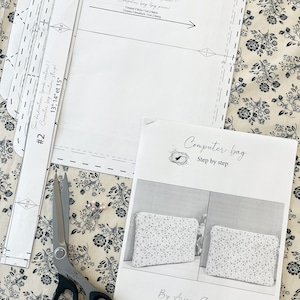 May include: A sewing pattern for a computer bag, with instructions in French and English. The pattern includes two pieces, one for the main bag and one for the lining. The pattern is laid out on a floral fabric with a pair of scissors and a few pins.