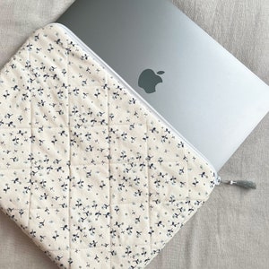 May include: A quilted laptop sleeve with a zipper closure and a tassel pull. The sleeve is made of a cream-colored fabric with a small floral pattern in blue and green.