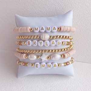 Peut inclure: Un ensemble de six bracelets avec des perles dorées et des perles blanches avec des lettres dorées épellant les noms "Mama", "Skylar" et "Emma".