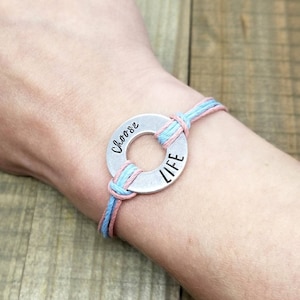 May include: A bracelet with a silver metal charm that says "Choose Life" on a light blue and pink cord.