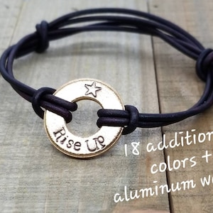 Peut inclure: Un bracelet en cuir noir avec un charme en métal doré qui dit "Rise Up" avec une étoile au-dessus du mot "Up".